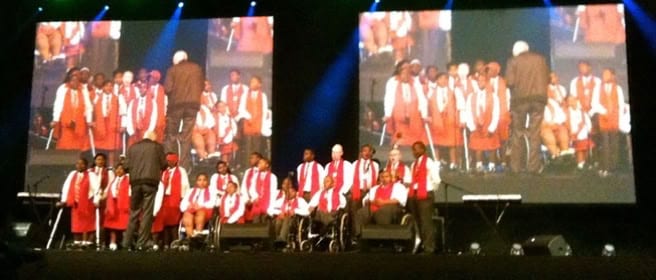 A choir performing on stage at a ceremony, shown on two large screens.