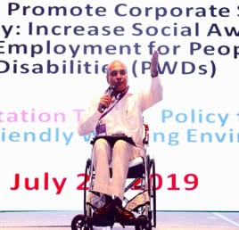 Abdus Sattar Dulal speaking from his wheelchair on stage in front of a banner about employment for persons with disabilities, July 2019.